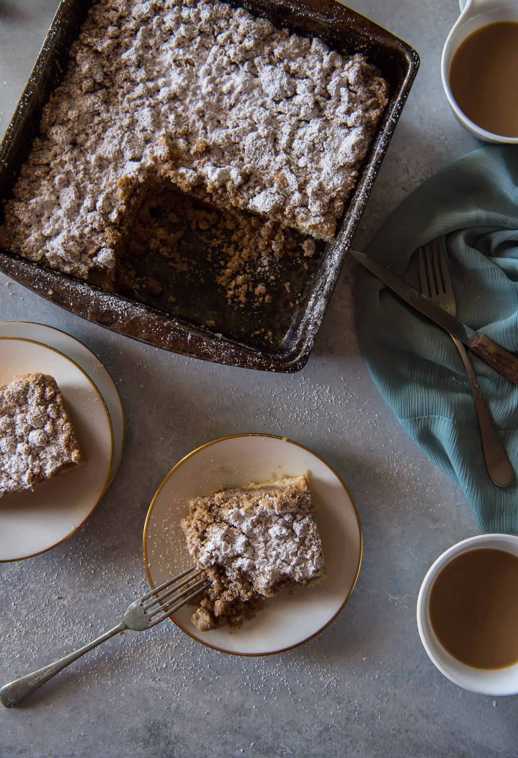an overhead photo of a pan of homemade crumb cake, plates with pieces of cake and a fork, and cups of coffee
