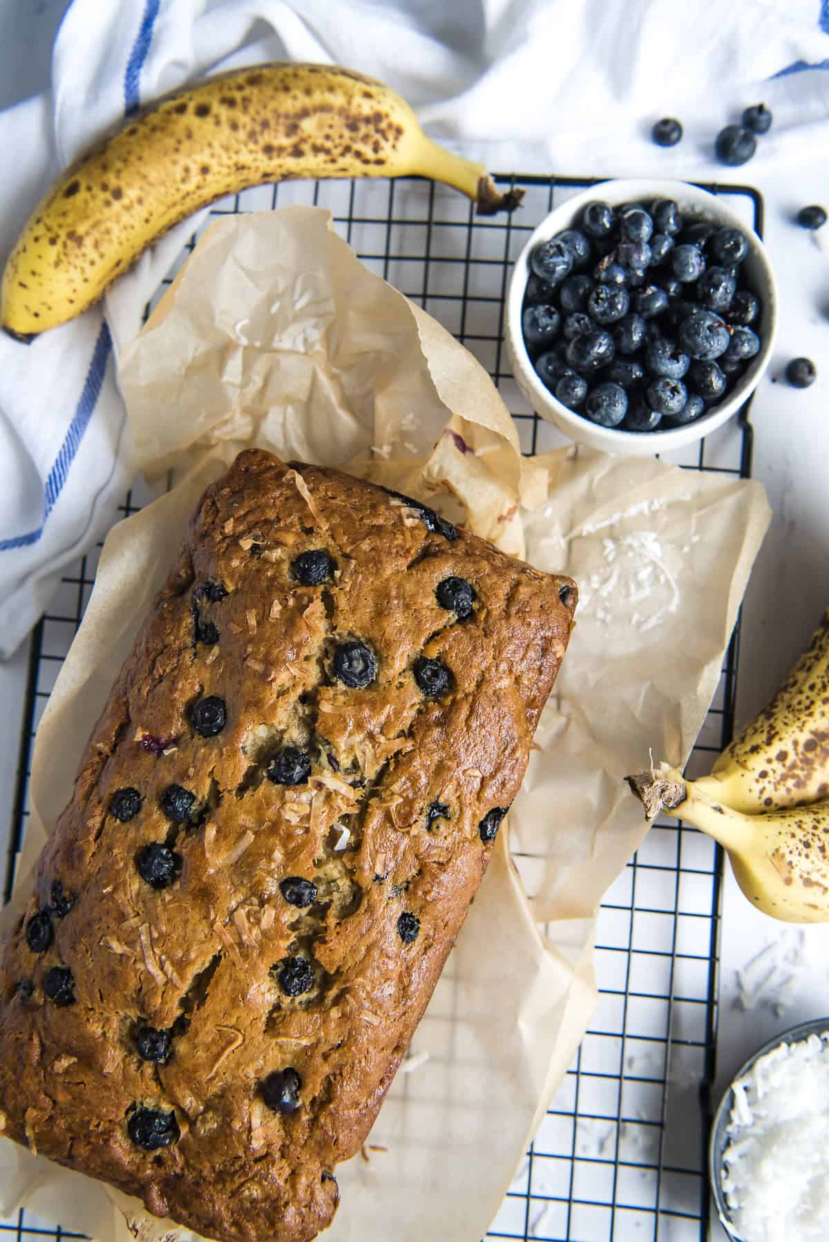 the top of a baked loaf of blueberry banana bread on a cooling rack with a bowl of blueberries and ripe bananas