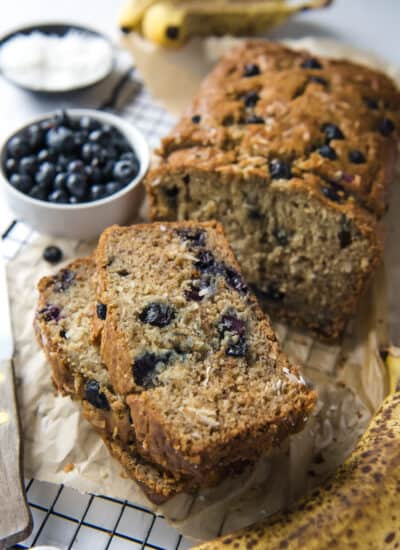 blueberry coconut banana bread slices on a cooling rack with bowls of coconut and blueberries and a banana in the background
