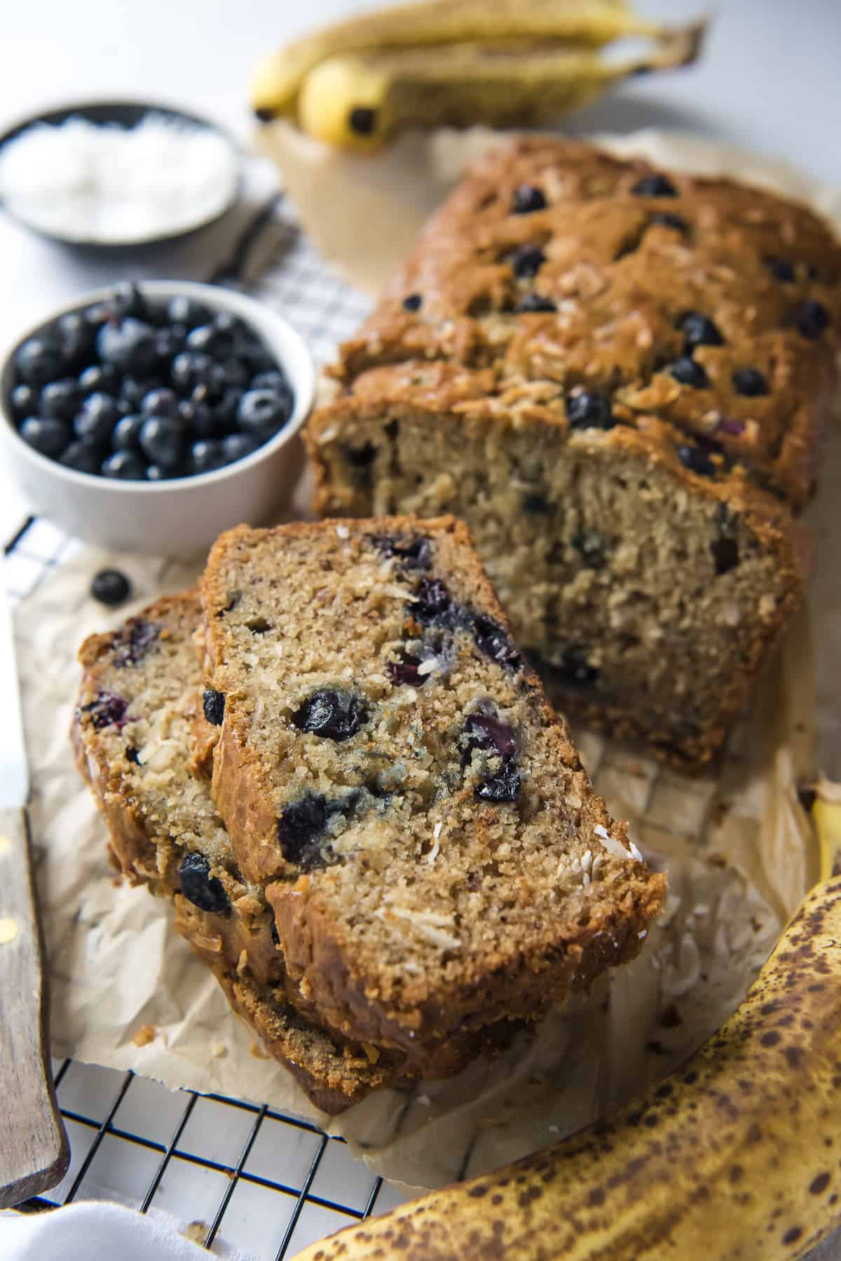 blueberry coconut banana bread slices on a cooling rack with bowls of coconut and blueberries and a banana in the background