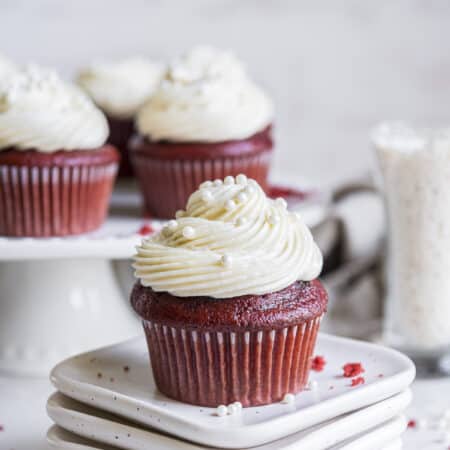 a red velvet cupcake on a stack of white plates with a cake stand full of more cupcakes in the background
