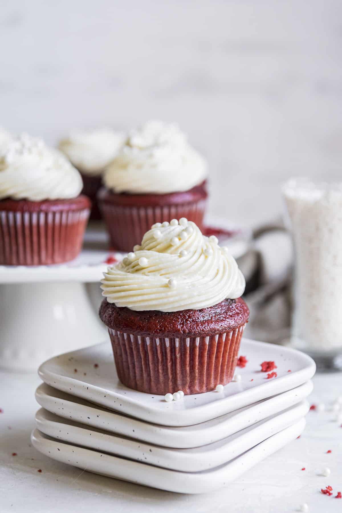 a red velvet cupcake on a stack of white plates with a cake stand full of more cupcakes in the background