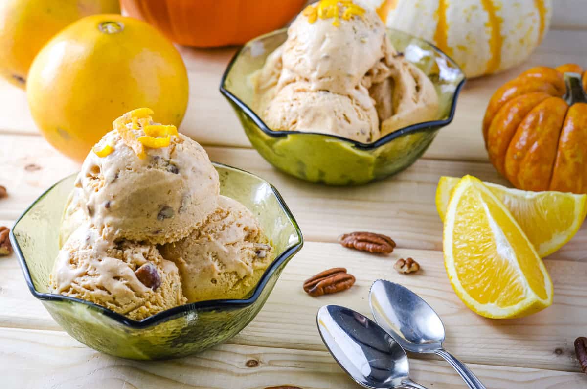 scoops of pumpkin ice cream in a green glass bowl surrounded by pecans, orange wedges, and spoons