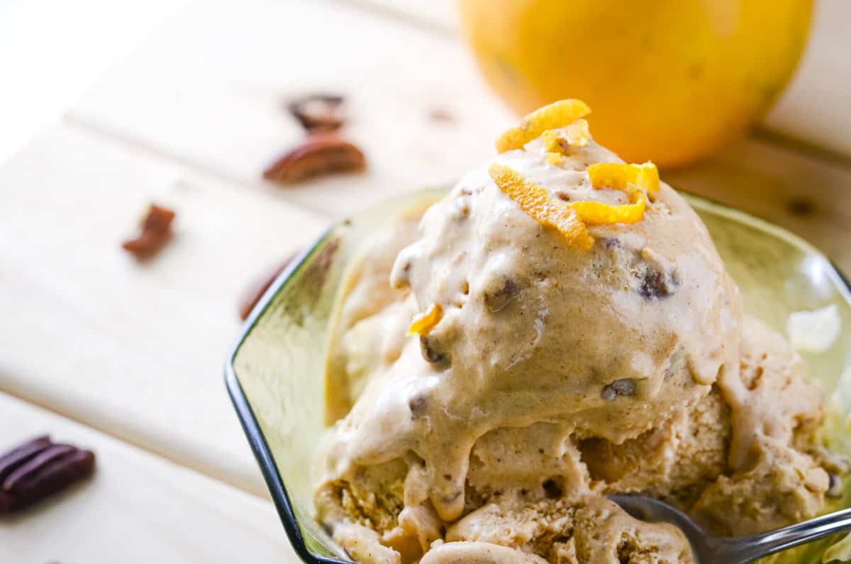a bowl of slightly melted pumpkin ice cream surrounded by pecans, orange wedges, and spoons