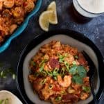 A bowl of slow cooker jambalaya with shrimp, sausage, rice, and vegetables, garnished with green onions and cilantro, served beside a lemon wedge and a glass of beer.