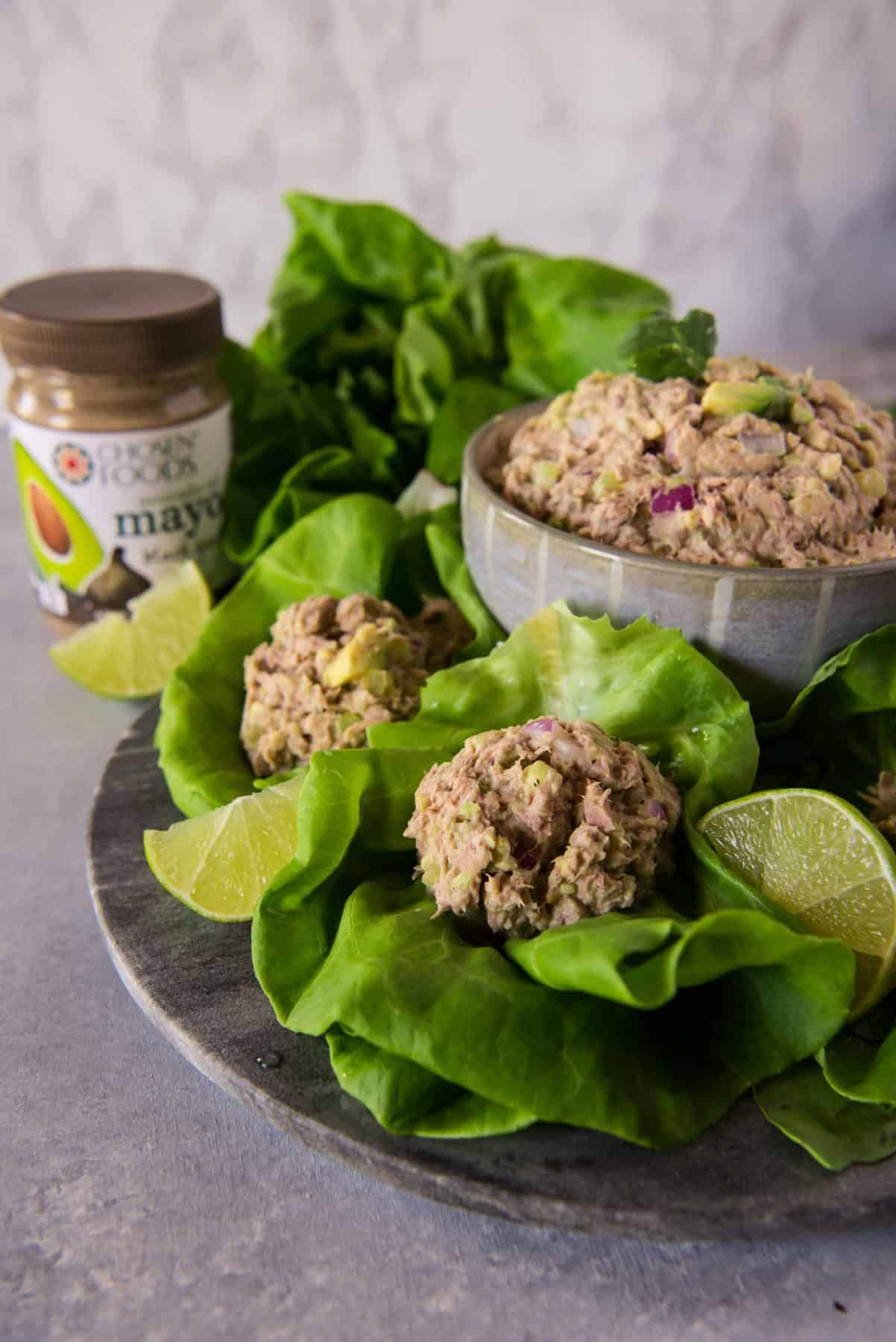 A bowl of avocado tuna salad served on lettuce leaves with lime slices, next to a jar of mayonnaise on a wooden surface.