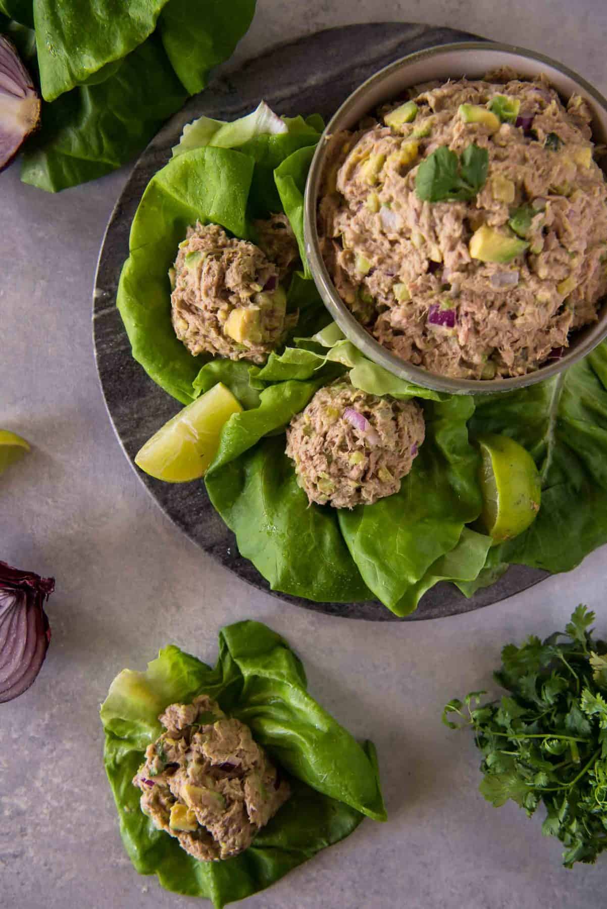 A bowl of avocado tuna salad with scoops on lettuce leaves, garnished with lime wedges and red onion, on a gray surface.