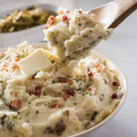 A bowl of creamy sour cream mashed potatoes garnished with chives, butter, and bacon bits, with a wooden spoon scooping out a portion.