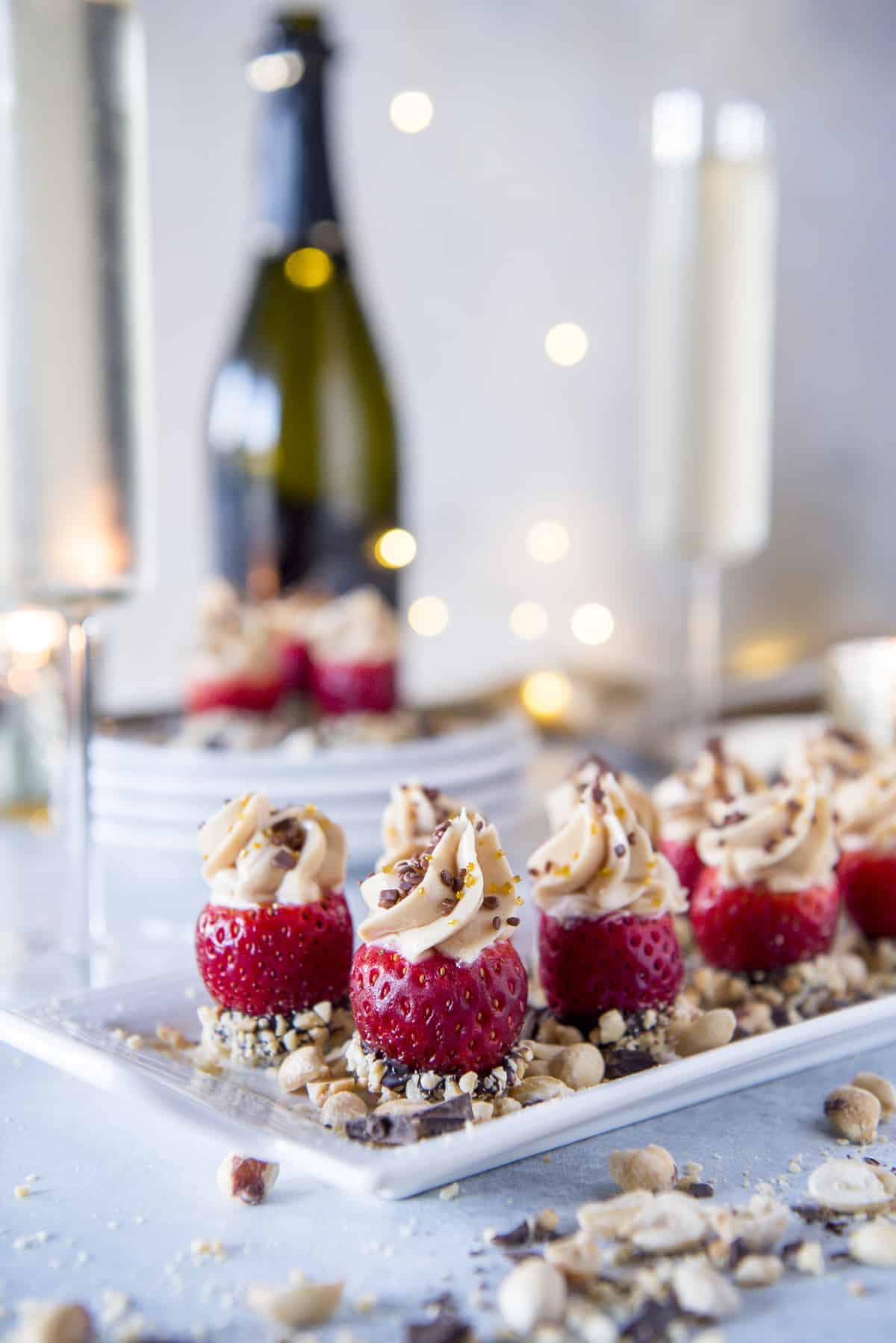 A plate topped with peanut butter cheesecake stuffed strawberries and nuts, with champagne glasses.