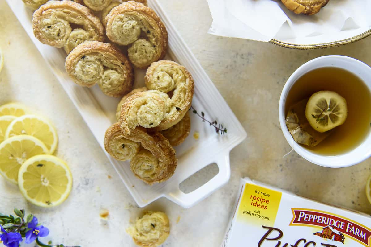 A top-down view of a table with a tray of flaky lemon thyme palmier cookies, a cup of tea with lemon, lemon slices, a box of Pepperidge Farm puff pastry