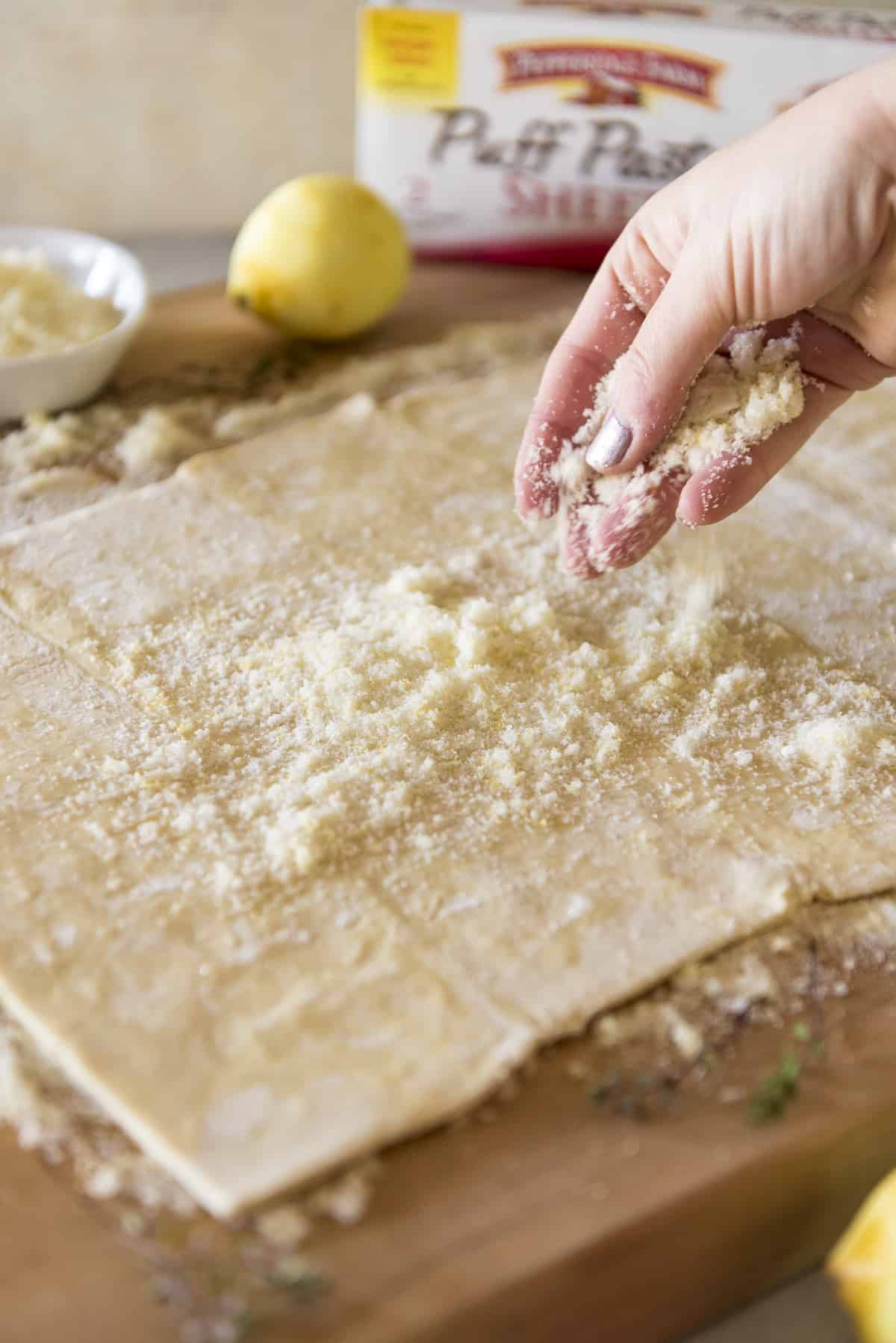 A person sprinkling sugar on a sheet of puff pastry on a wooden board, with a lemon in the background.
