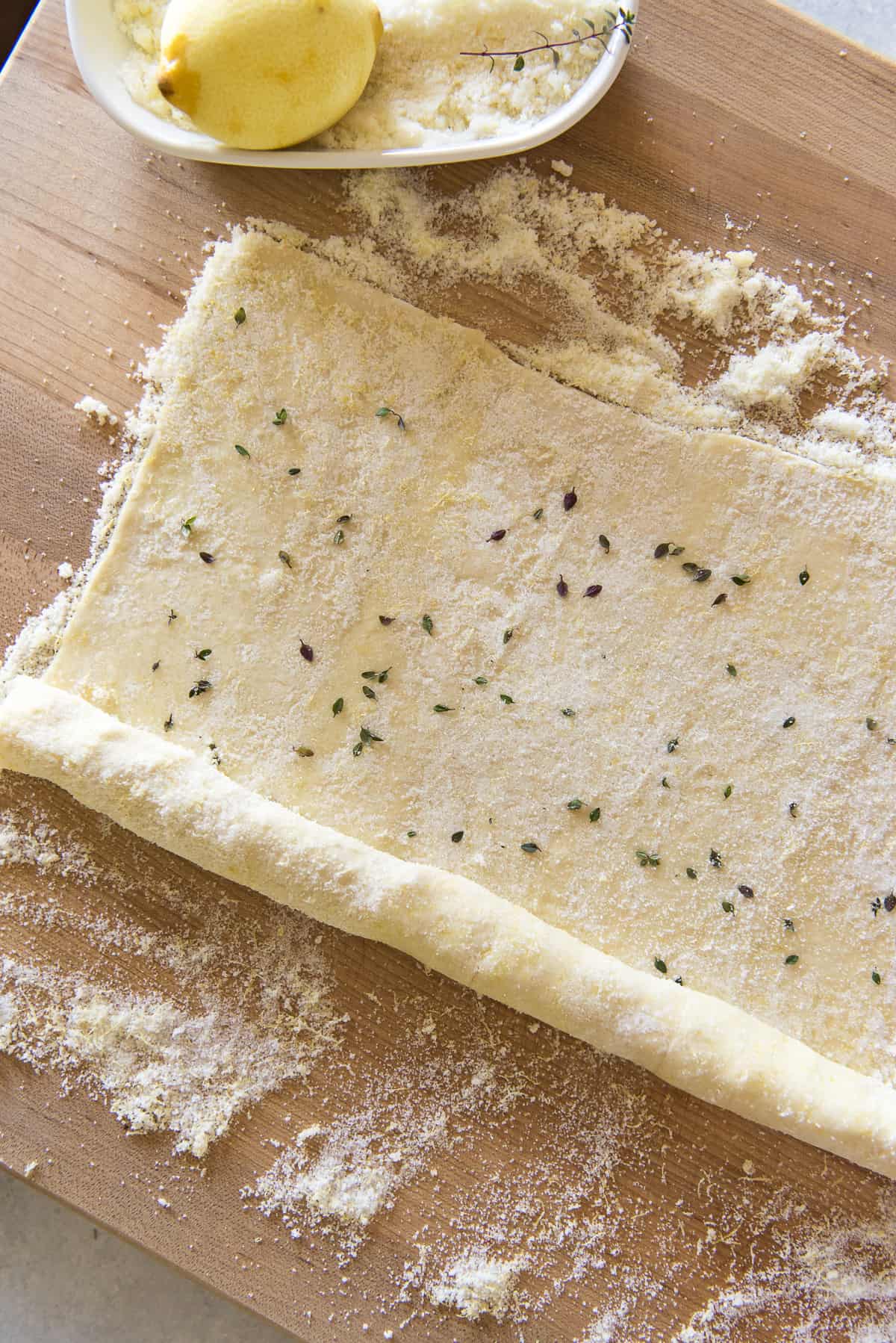 Uncooked puff pastry dough sprinkled with lemon sugar and thyme, rolled out on a wooden surface, with a bowl of filling nearby.