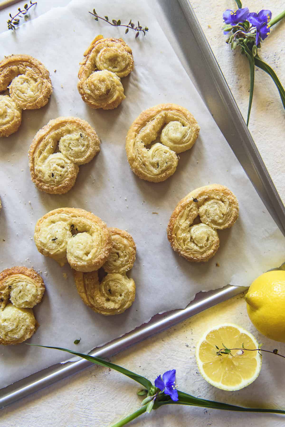 Freshly baked lemon palmiers pastry, arranged on a baking sheet with parchment paper, decorated with lemon slices and purple flowers.
