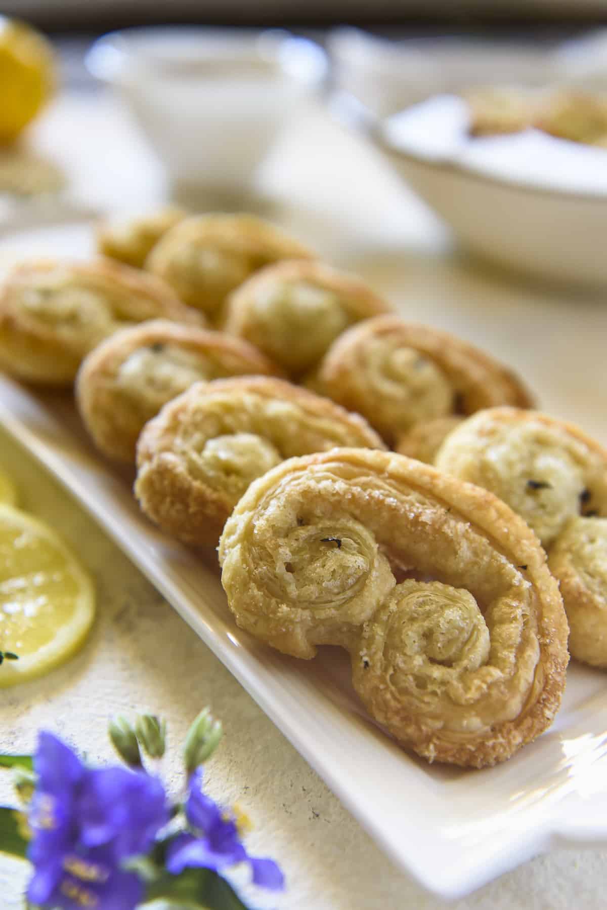 A platter of freshly baked lemon thyme palmiers garnished with lemon slices and flowers on a light-colored table.