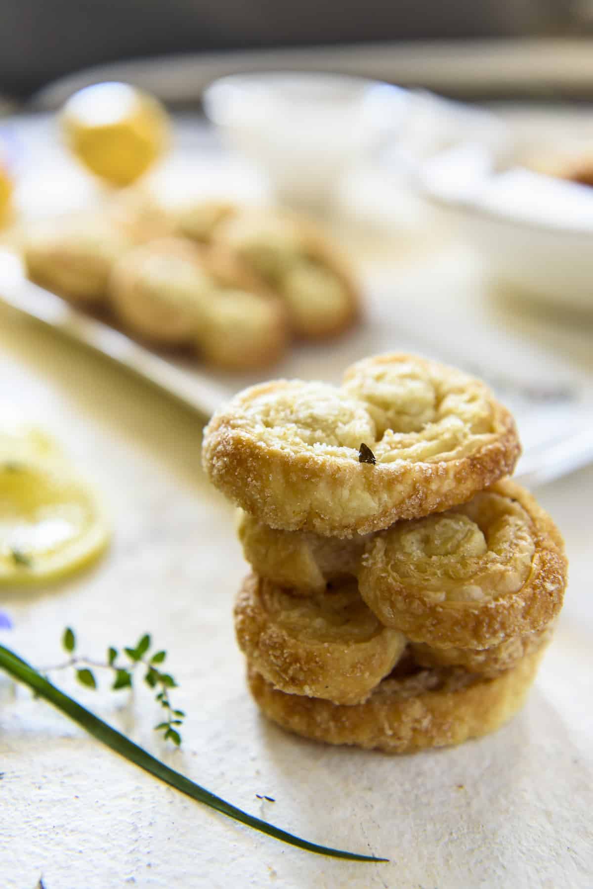 A stack of four flaky, golden-brown lemon palmiers on a white surface, with blurred cookies and small dishes in the background.