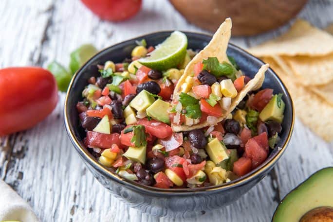 A bowl of Avocado Black Bean and Corn Salsa with a tortilla chip dipped inside