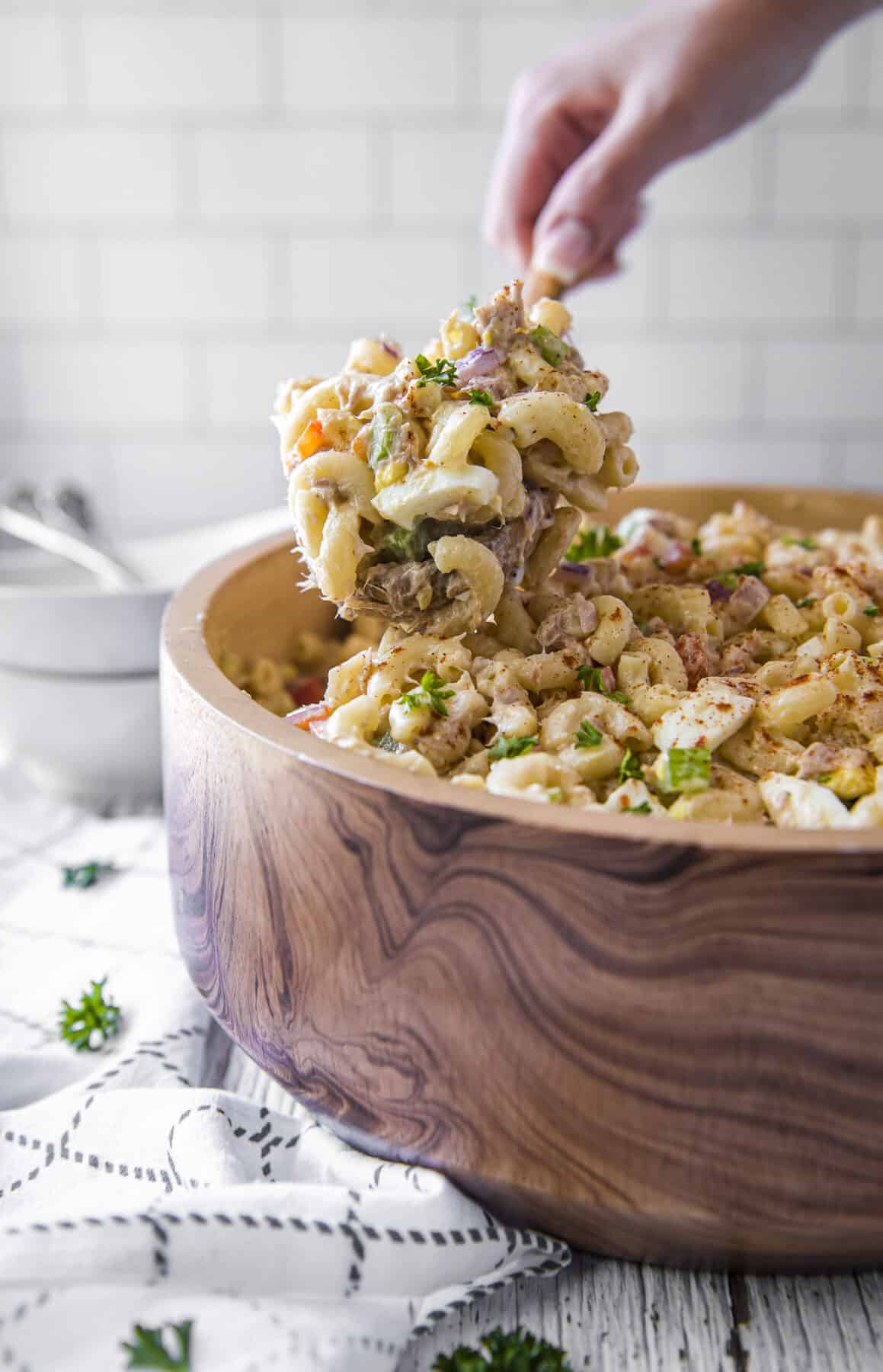 A hand lifting a scoop of tuna pasta salad from a wooden bowl
