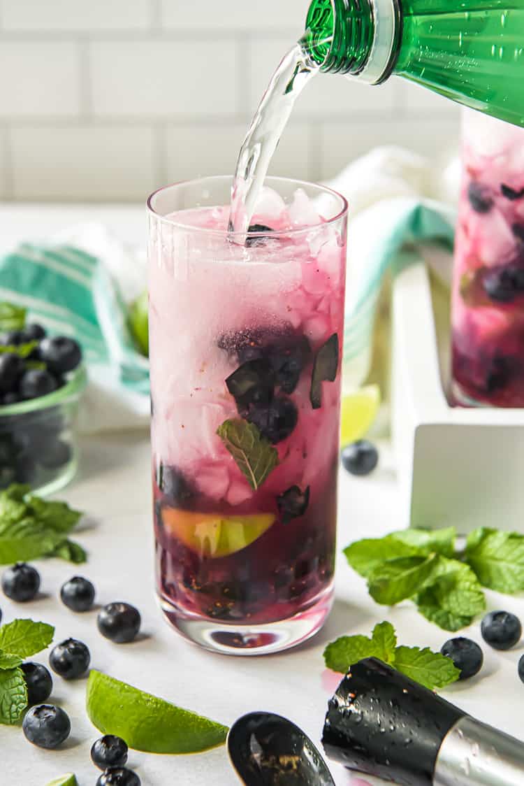 pouring sparkling water into a blueberry mojito