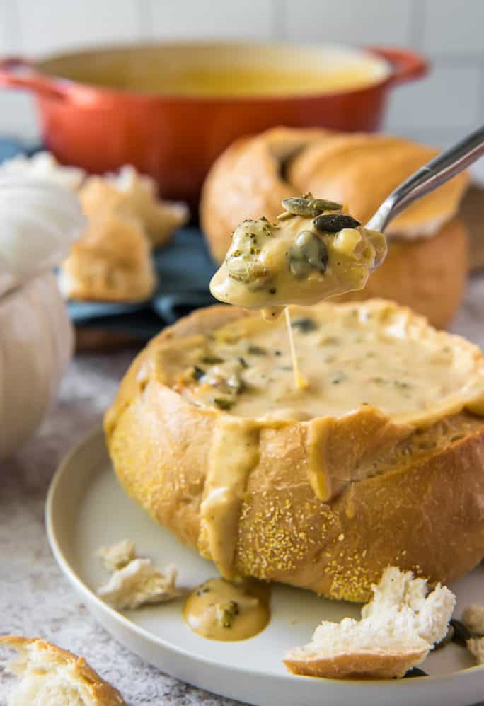 a spoon lifting pumpkin broccoli cheddar soup from a sourdough bread bowl