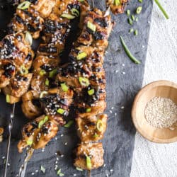 a pile of grilled teriyaki chicken skewers on a slate platter with a bowl of sesame seeds on the side