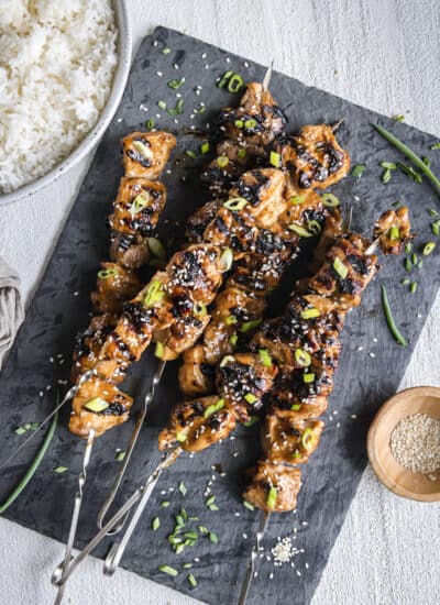 a pile of grilled teriyaki chicken skewers on a slate platter with a bowl of rice and sesame seeds on the side