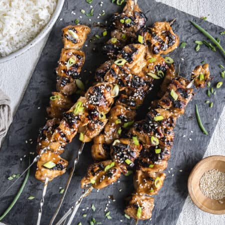a pile of grilled teriyaki chicken skewers on a slate platter with a bowl of rice and sesame seeds on the side