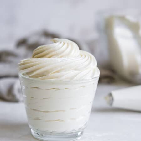 whipped cream cheese frosting piped into a small glass container in front of a piping bag
