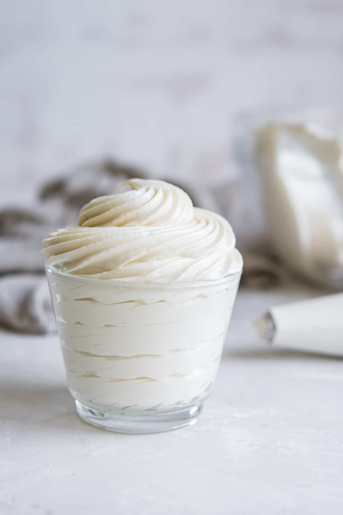 whipped cream cheese frosting piped into a small glass container in front of a piping bag
