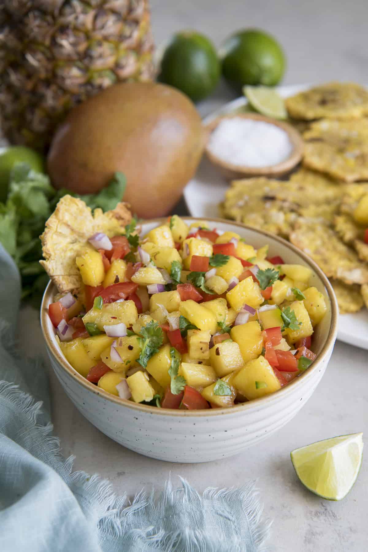 A fried tostone dipped in a white bowl of Pineapple Mango Salsa, surrounded by cilantro, limes, mango, a pineapple, and a platter of fried tostones