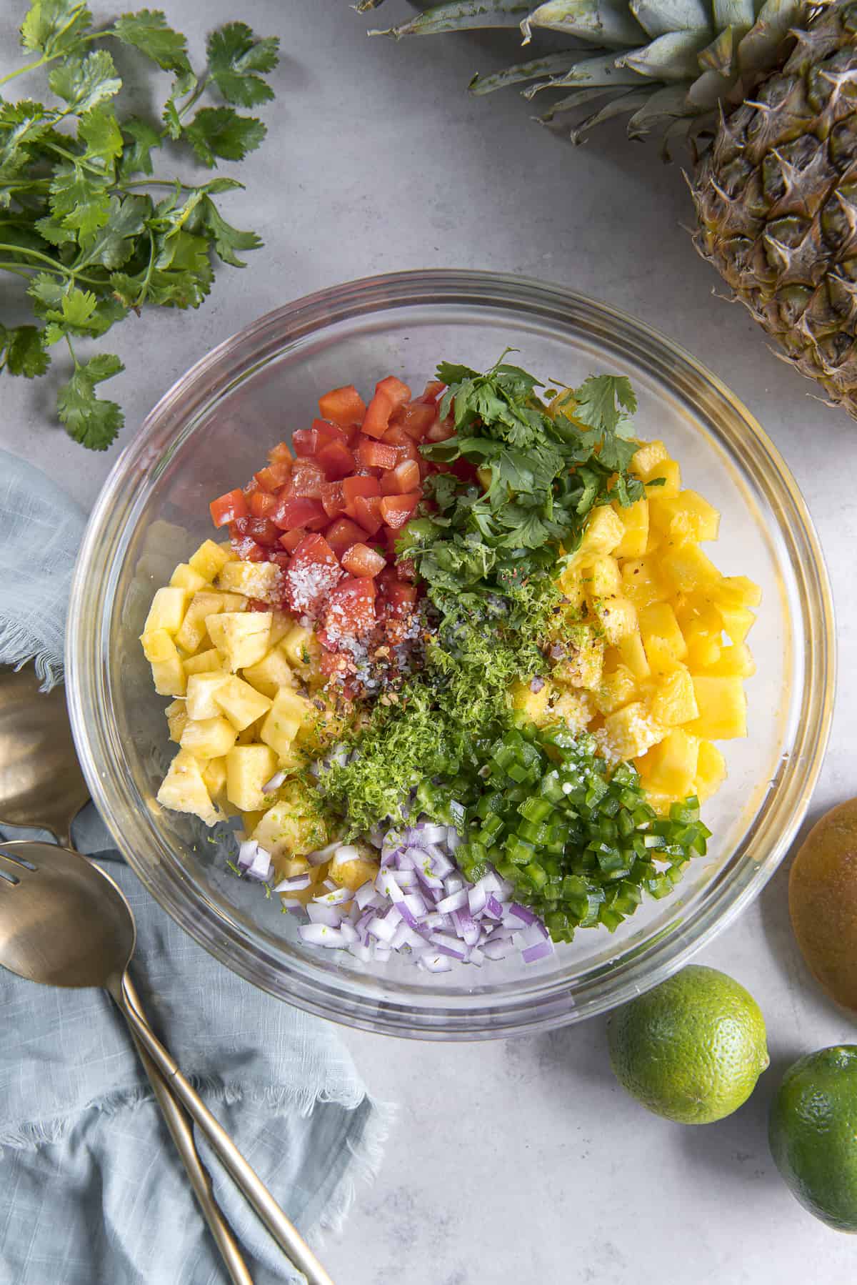 the ingredients for pineapple mango salsa in a glass mixing bowl, surrounded by limes, cilantro, mango, a pineapple, and gold serving spoons on top of a blue napkin