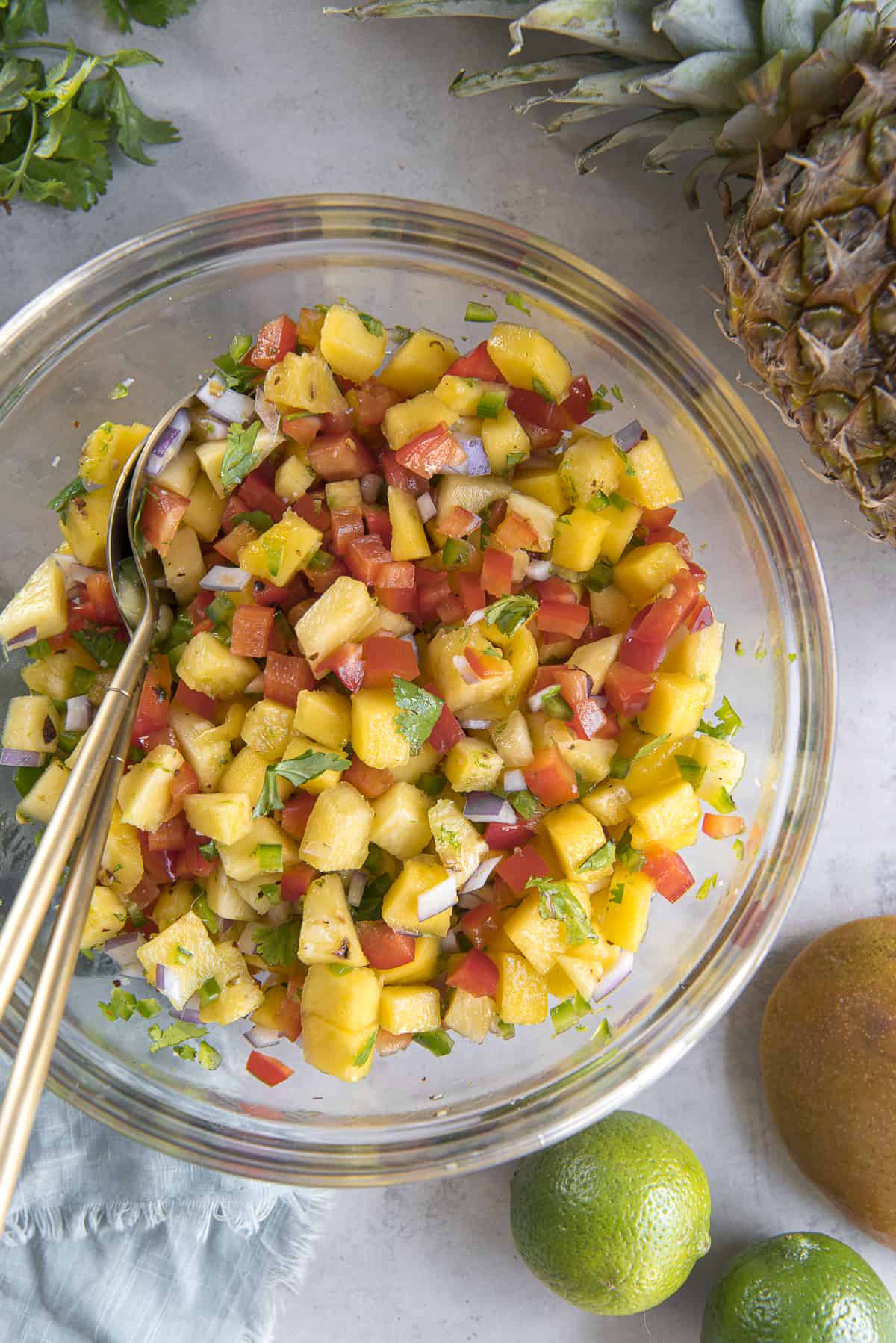the ingredients for pineapple mango salsa tossed together in a glass mixing bowl with gold utensils, surrounded by limes, cilantro, mango, a pineapple, and a blue napkin