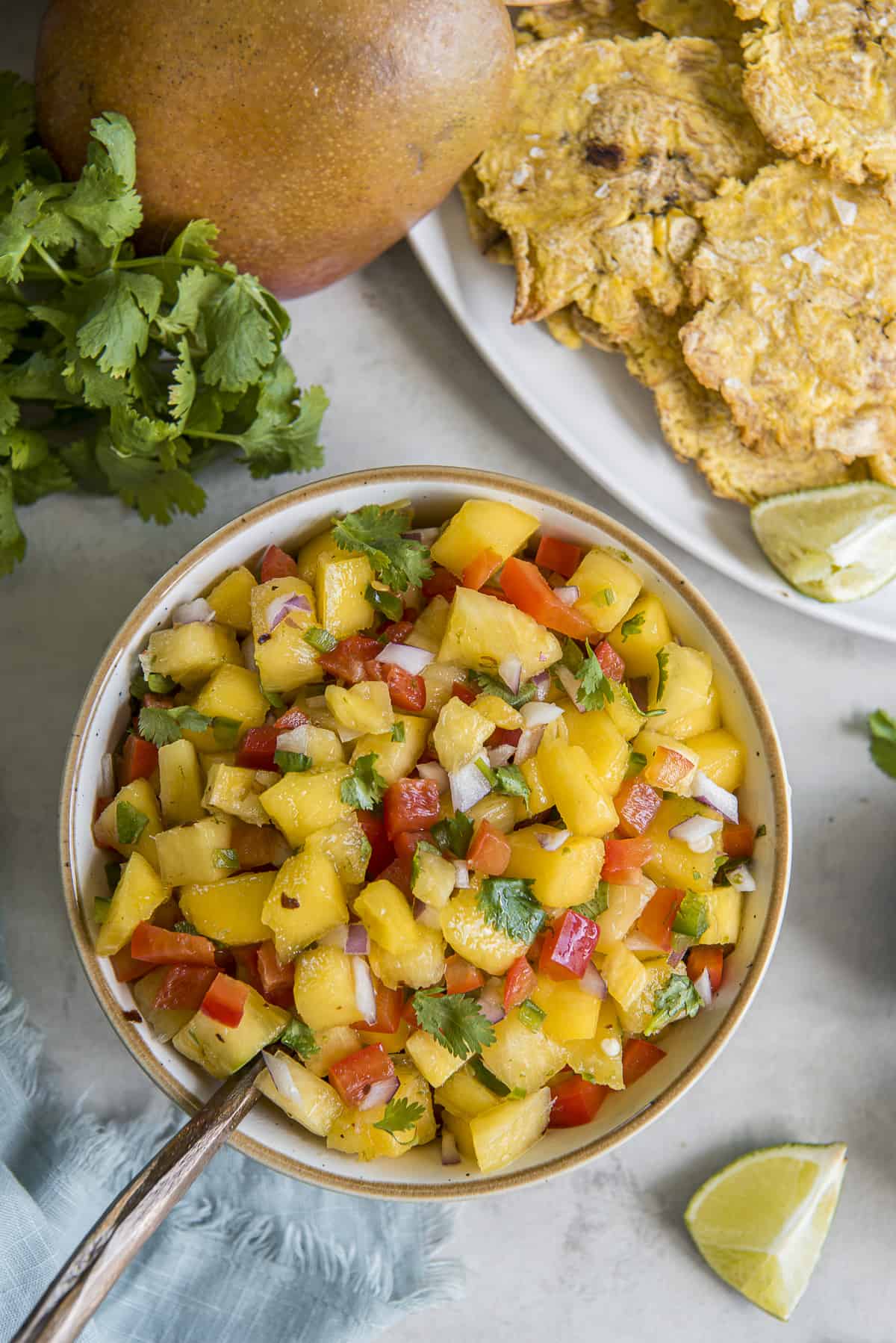 An overhead shot of Pineapple Mango Salsa in a white bowl with a spoon, surrounded by cilantro, limes, mango, a pineapple, and a platter of fried tostones