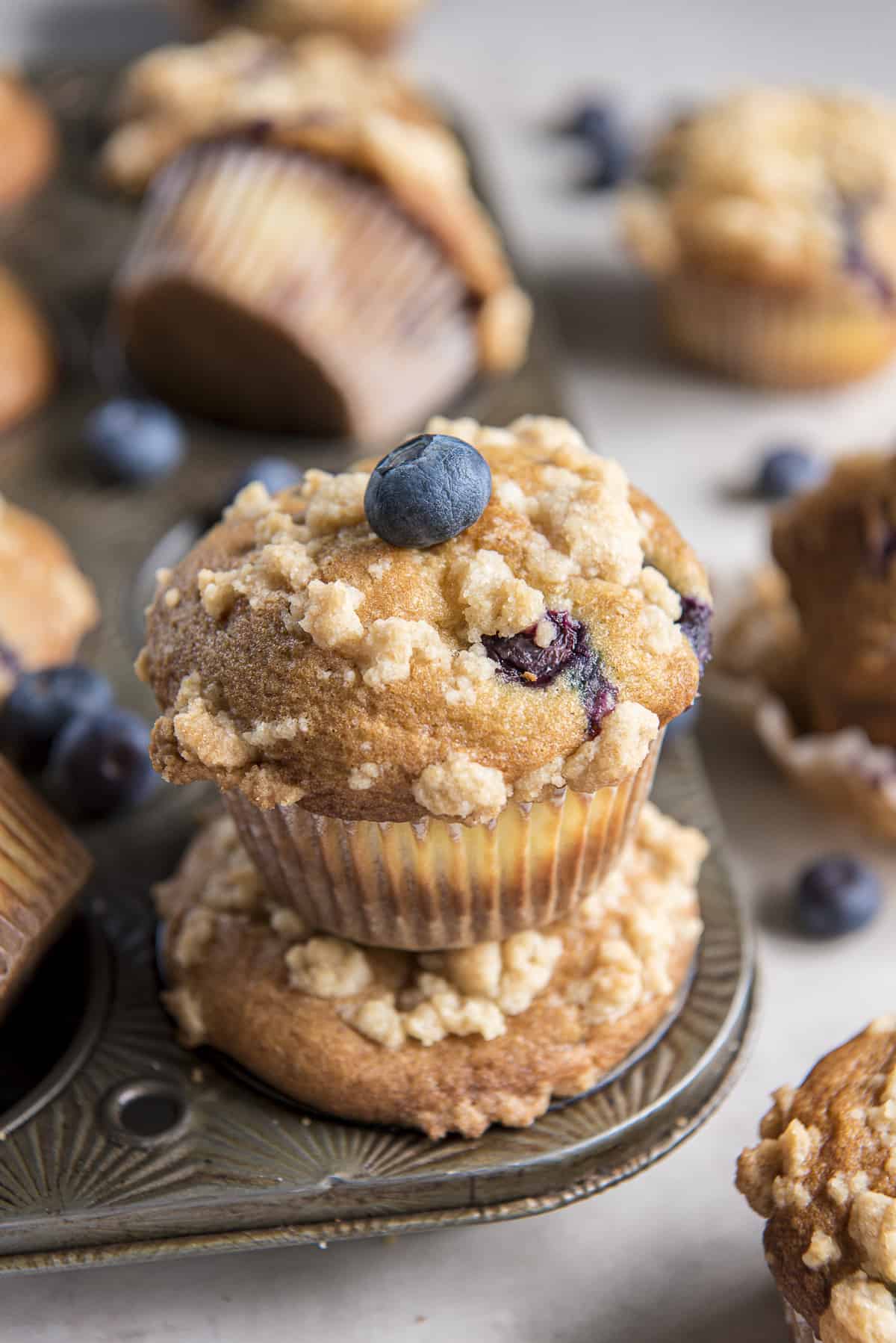 two blueberry cream cheese muffins stacked on top of each other in a muffin tin, with several more scattered around the background