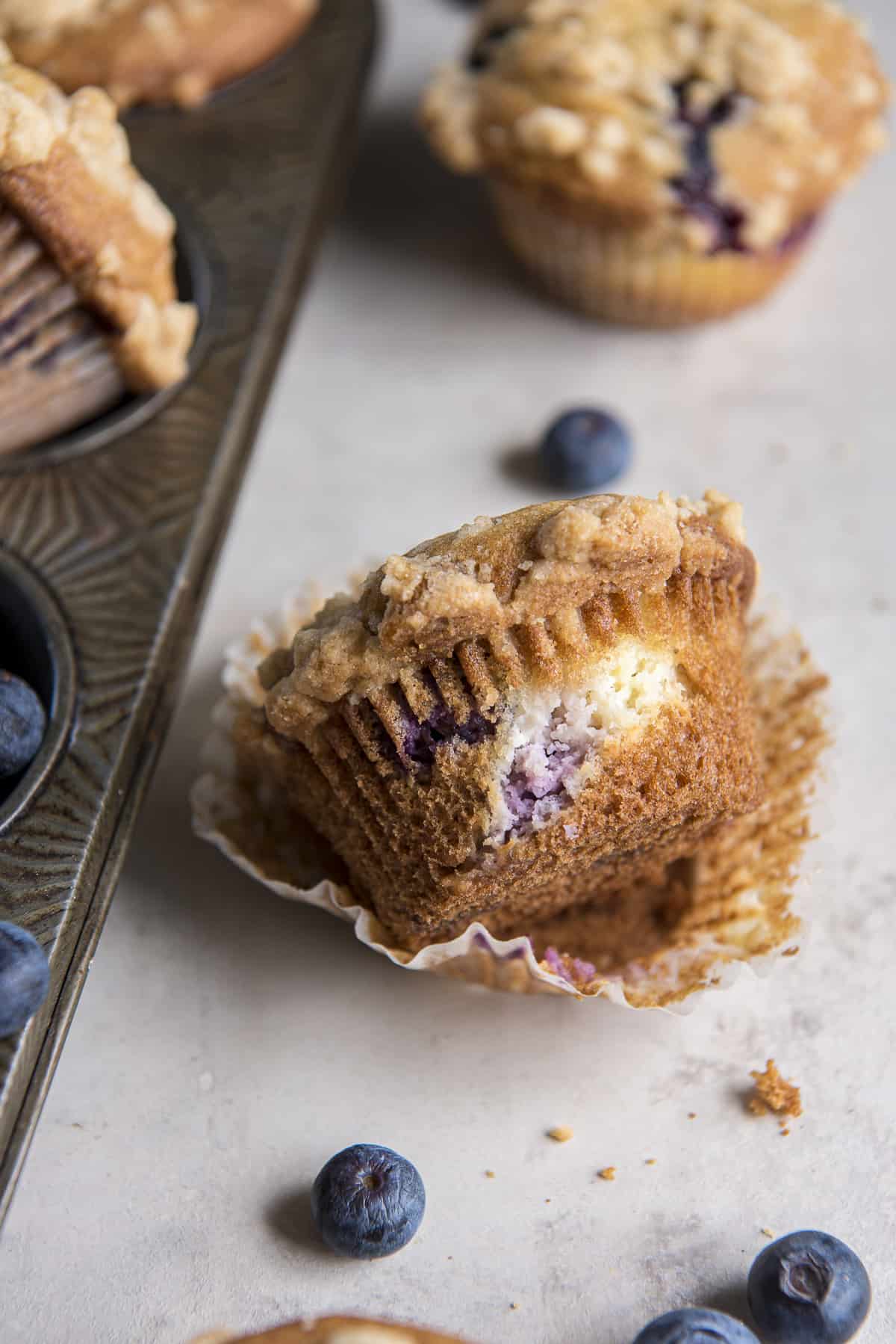 an unwrapped blueberry cream cheese muffin next to a muffins tin