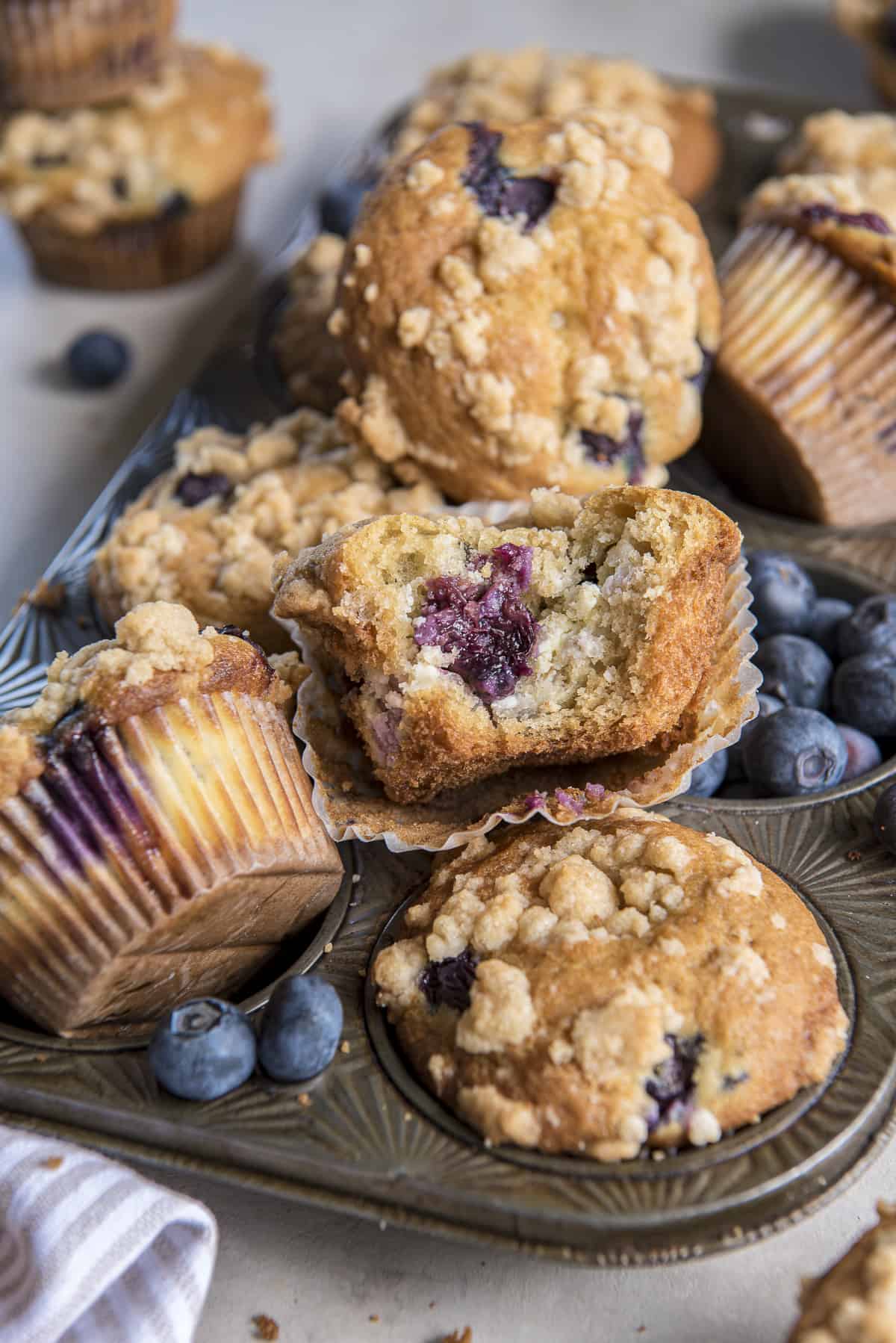 a blueberry cheesecake muffin with a bite taken out of it, nestled among other muffins in a vintage muffin tin with blueberries scattered around