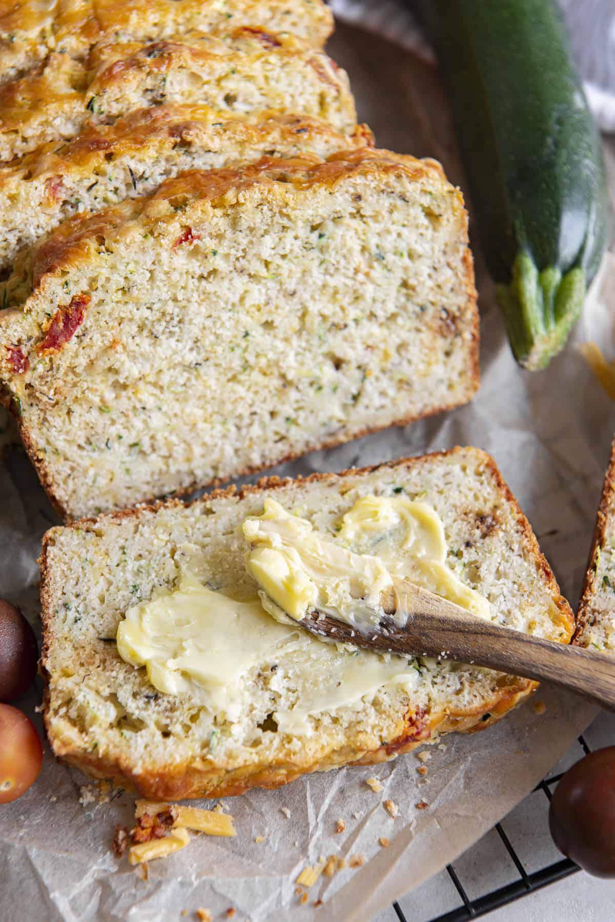 a wooden knife spreading butter on a piece of savory zucchini bread sitting on parchment, surrounded by vegetables