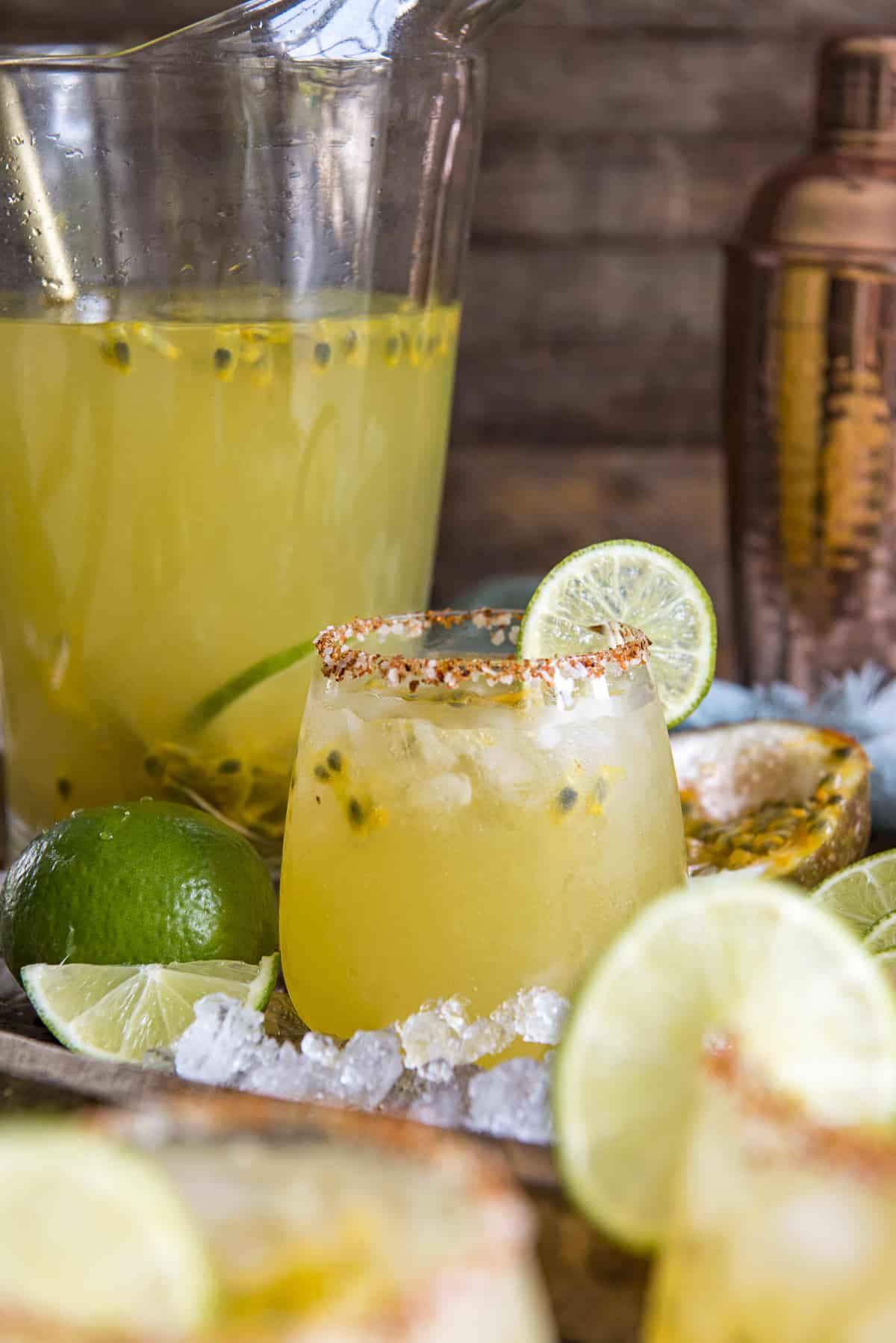 a glass of passion fruit margarita on a wooden tray with a pitcher of margaritas behind