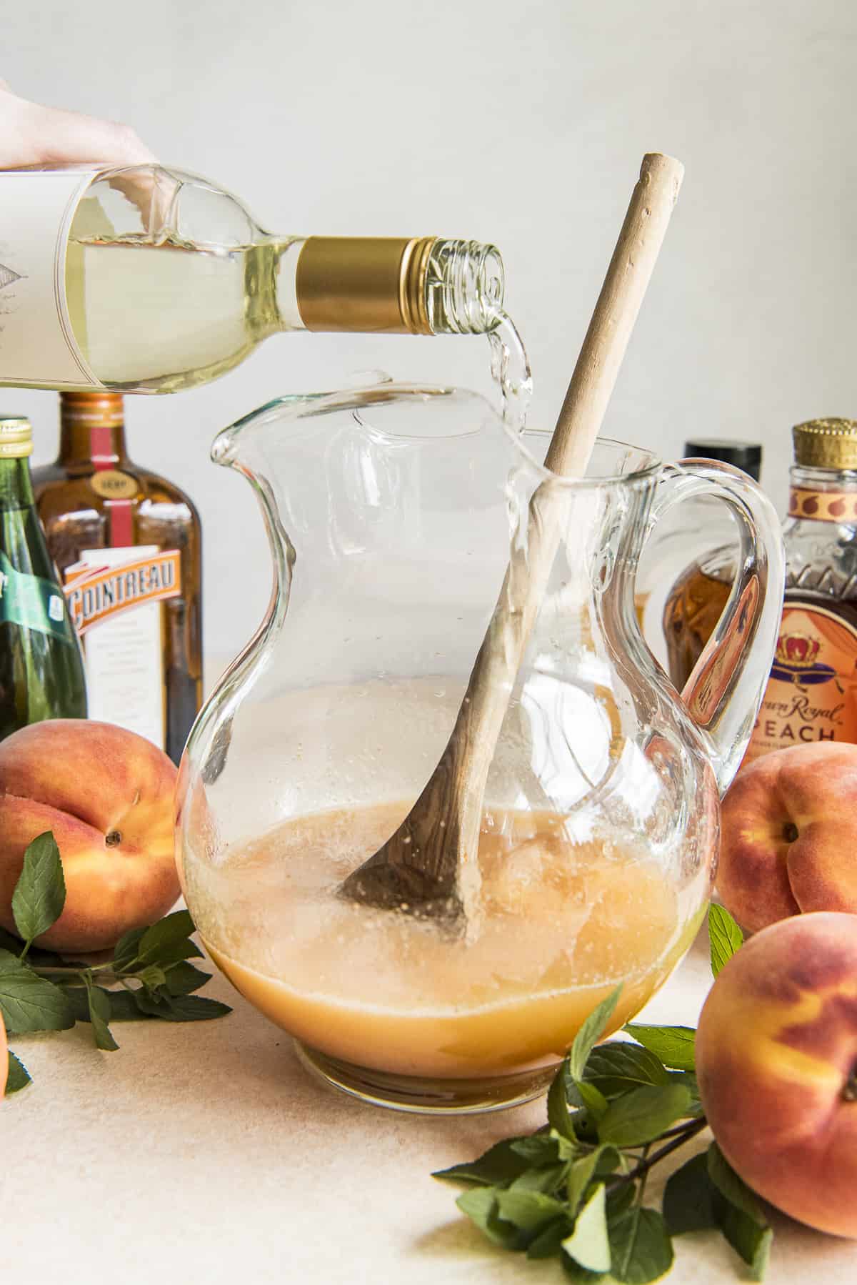 a hand pouring a bottle of white wine into a pitcher of peach nectar