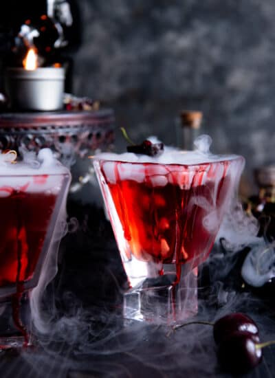 Two glasses of red vampire's kiss cocktail with smoke and cherries in them.