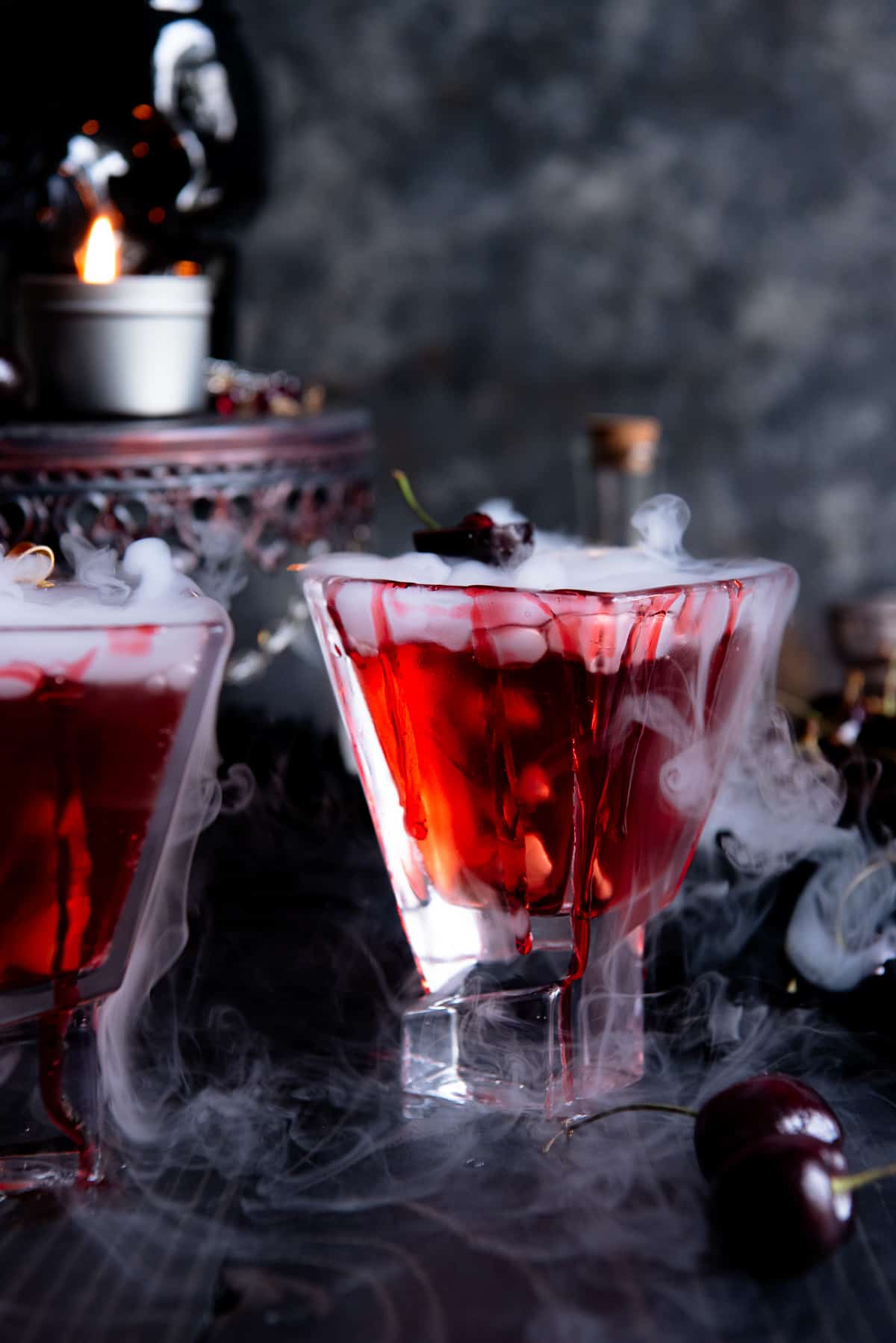 Two glasses of red liquid with smoke and cherries in them.